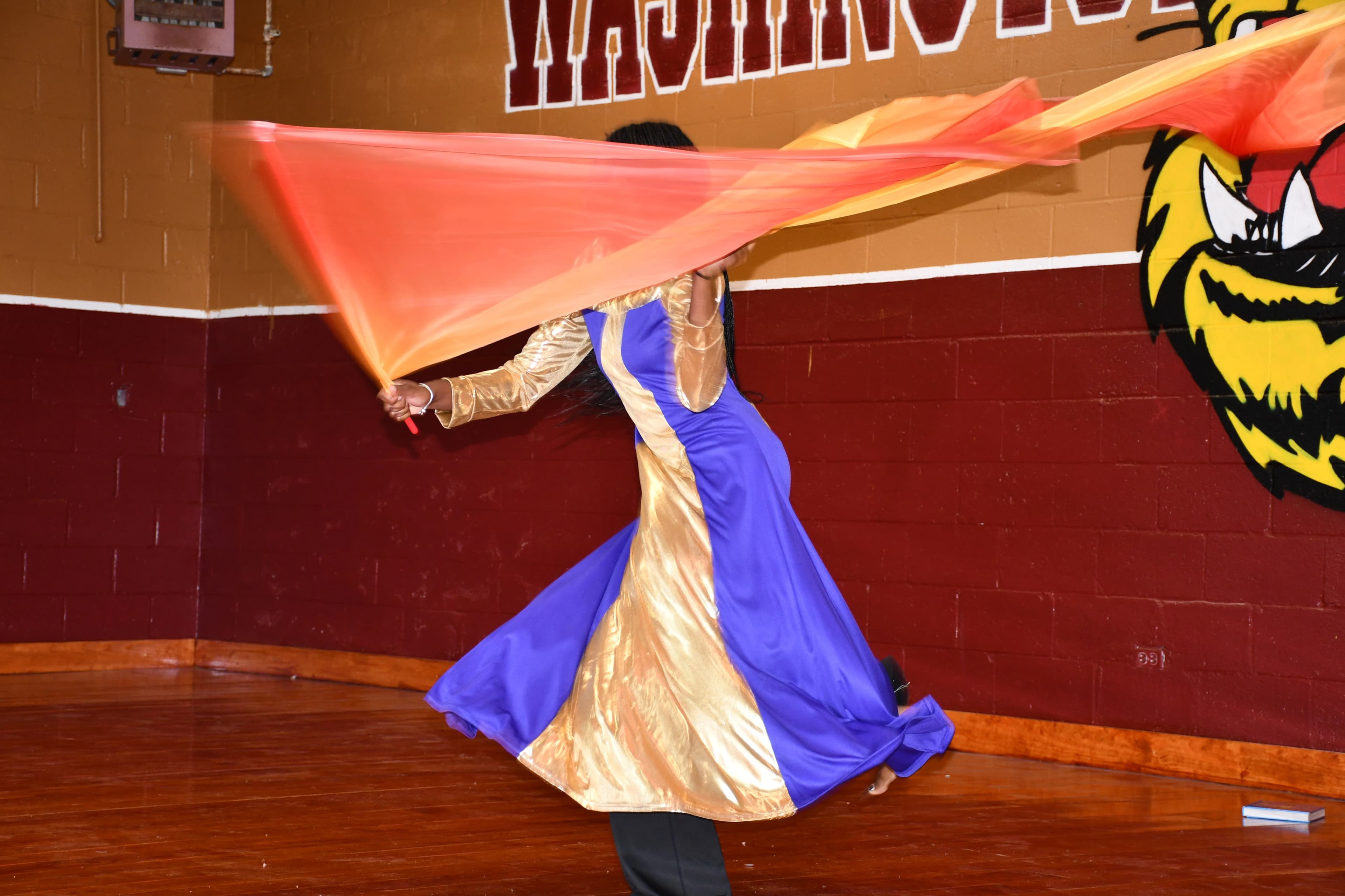 A praise dancer in a blue and gold robe waving worship flags beneath the Washington High School tiger mural